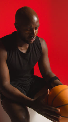 Vertical-Video-Close-Up-Studio-Shot-Of-Seated-Male-Basketball-Player-With-Hands-Holding-Ball-Against-Red-Background-1